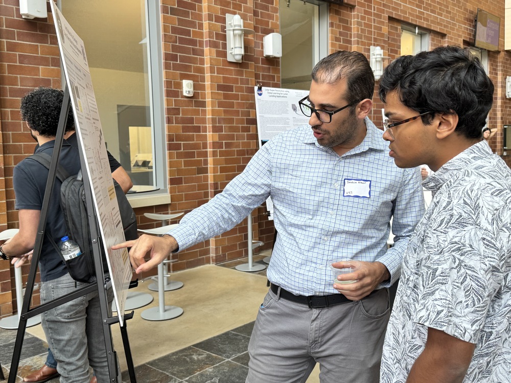 Two men standing near a poster, with one pointing at the poster.